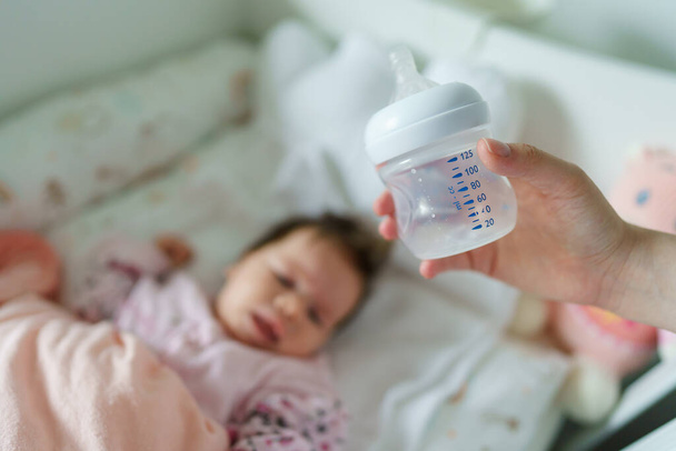 Close up selective focus on empty baby bottle in hands of unknown woman mother holding it above the bed at home with sleeping infant in background - newborn feeding nursing concept concept copy space - Fotografie, Obrázek