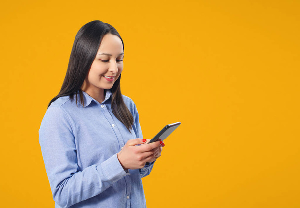 Mujer joven usando smartphone y sonriendo. Sobre un fondo amarillo. - Foto, Imagen
