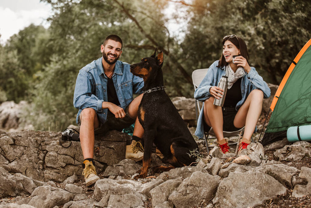 Man and woman and their doberman sitting in chairs outside the tent having coffee and talking. Couple camping. - Photo, Image