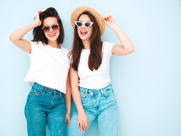 Two young beautiful smiling hipster female in trendy summer white t-shirt and jeans clothes.Sexy carefree women posing near light blue wall in studio.Positive models in hats - 写真・画像