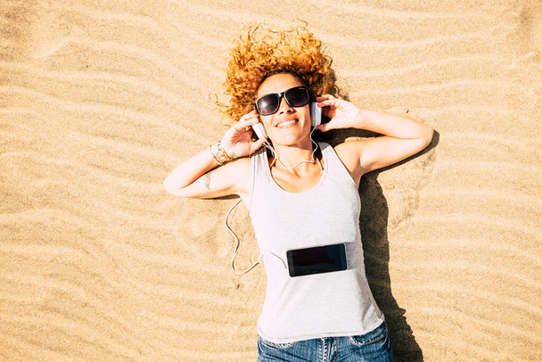 Cheerful happy woman lay down at the beach listening music with phone and headphones enjoying summer travel holiday vacation alone in top view  - Фото, зображення
