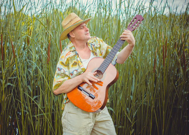 Hombre mayor divertido en camisa estampada turista tocando la guitarra y cantar canción en la naturaleza al aire libre, viaje a América y placer para las vacaciones - Foto, Imagen