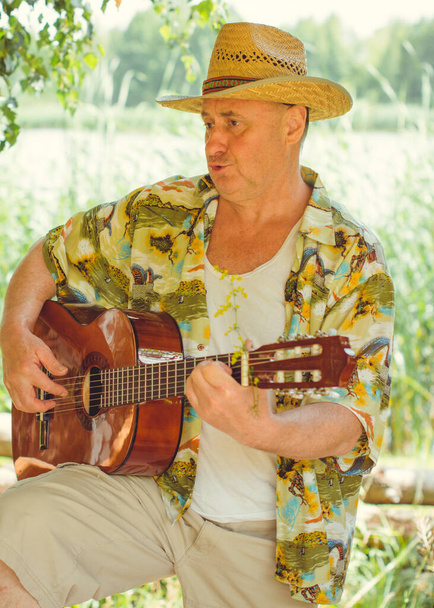 Смешной пожилой человек в печатной рубашке турист, играющий на гитаре и поющий песню на природе на открытом воздухе, поездка в Америку и удовольствие для отдыха - Фото, изображение