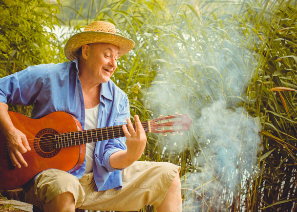 Homem mais velho engraçado na camisa de impressão turista tocando guitarra e cantar música na natureza ao ar livre, viagem para a América e prazer para férias - Foto, Imagem