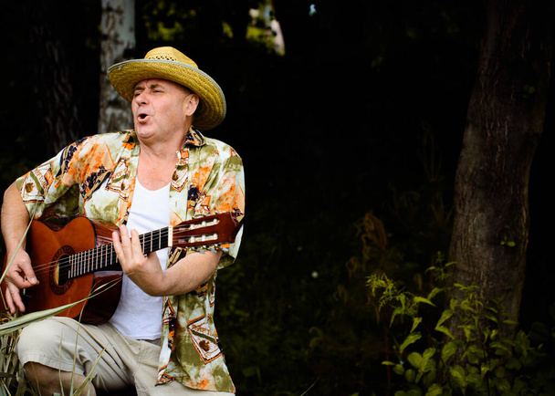 Funny older man in print shirt tourist playing guitar and sing song on nature outdoors, trip to America and pleasure for vacation - Fotoğraf, Görsel