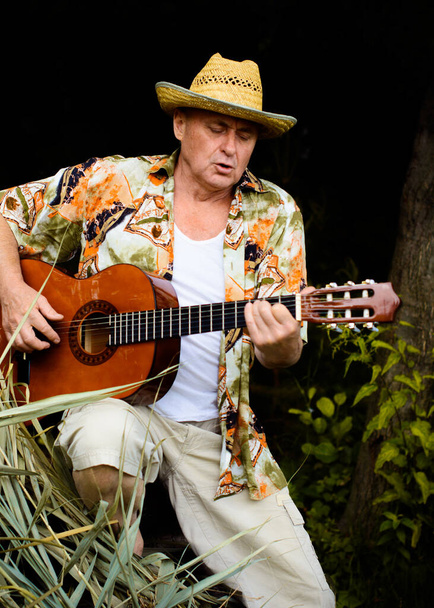 Αστείος ηλικιωμένος άνδρας σε έντυπη φανέλα τουρίστας παίζει κιθάρα και τραγουδούν τραγούδι στη φύση σε εξωτερικούς χώρους, το ταξίδι στην Αμερική και την ευχαρίστηση για διακοπές - Φωτογραφία, εικόνα