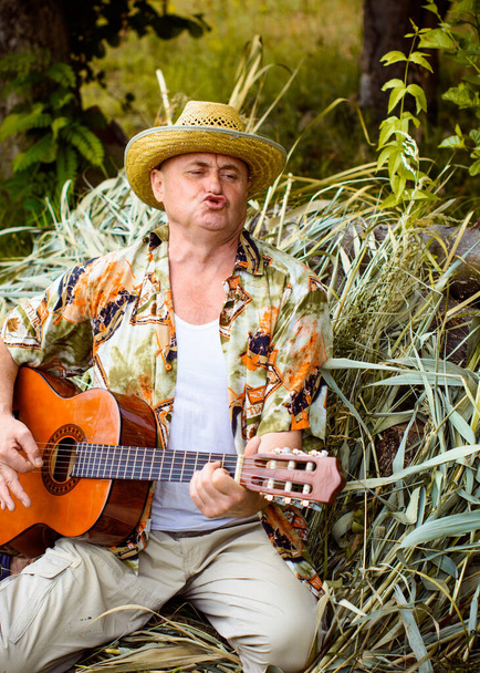 Αστείος ηλικιωμένος άνδρας σε έντυπη φανέλα τουρίστας παίζει κιθάρα και τραγουδούν τραγούδι στη φύση σε εξωτερικούς χώρους, το ταξίδι στην Αμερική και την ευχαρίστηση για διακοπές - Φωτογραφία, εικόνα