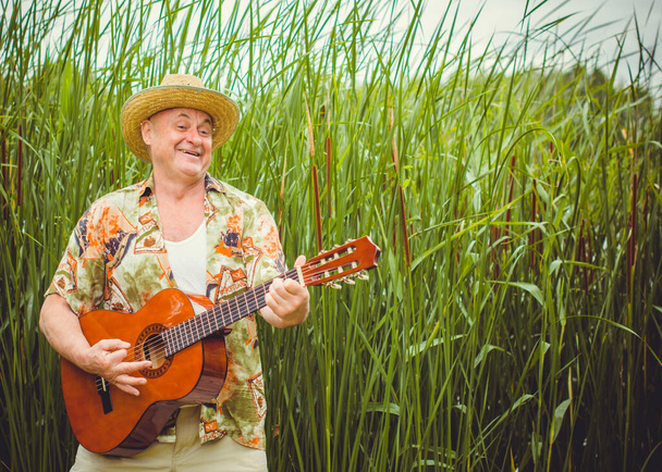 Funny older man in print shirt tourist playing guitar and sing song on nature outdoors, trip to America and pleasure for vacation - Photo, Image