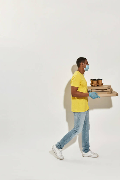 Full length shot of delivery man wearing protective mask and gloves looking away, carrying pizza boxes and drinks isolated over gray background - Foto, imagen