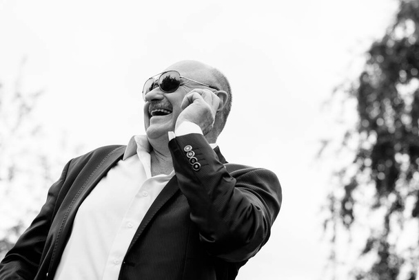Portrait stylischer reifer Männer in weißem Hemd und Sakko und blauer Sonnenbrille per Smartphone. Mit seinem Gerät bedient er das Internet. Alter Mann mit sozialer Netzwerktechnologie. Senior telefoniert - Foto, Bild