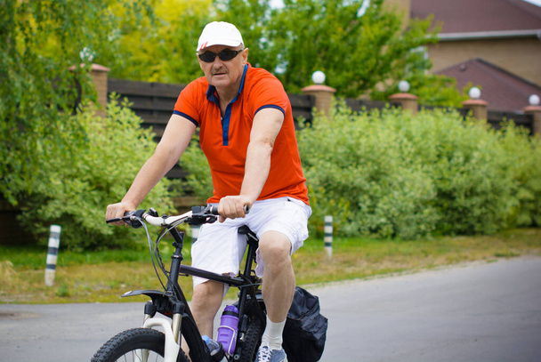 Ένας ώριμος άντρας με σορτς, ένα μπλουζάκι και ένα καπέλο κάνει βόλτες με το ποδήλατο στην εξοχική πόλη. Η έννοια του υγιεινού τρόπου ζωής μετά από 50 χρόνια - Φωτογραφία, εικόνα