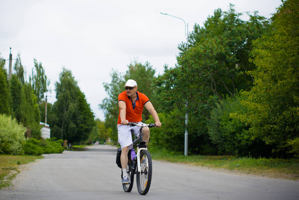 A mature man in shorts, a T-shirt and a cap rides a bicycle around the cottage town. The concept of a healthy lifestyle after 50 years - Photo, Image