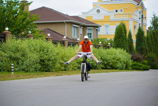 Şortlu, tişörtlü ve şapkalı olgun bir adam kır evi kasabasında bisiklet sürüyor. 50 yıl sonra sağlıklı yaşam biçimi kavramı - Fotoğraf, Görsel