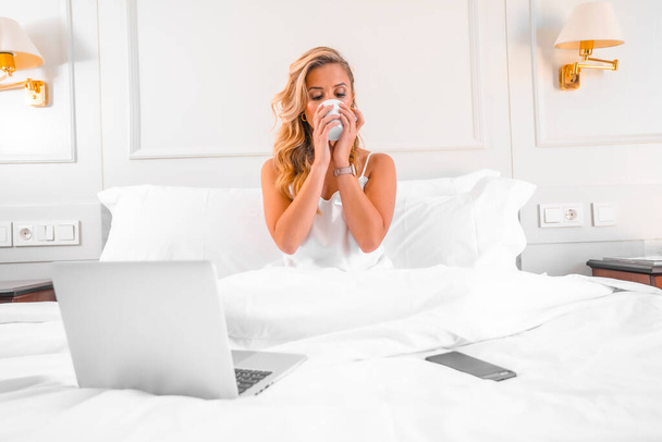 Portrait attractive young blond Caucasian woman in nightgown in hotel bed teleworking with computer. Working and traveling in a hotel - 写真・画像