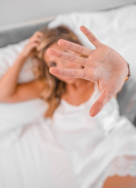 Portrait of an attractive young blond Caucasian woman in nightgown on the bed, putting her hand to the camera. Lifestyle in the hotel bed, overhead shot - Foto, immagini