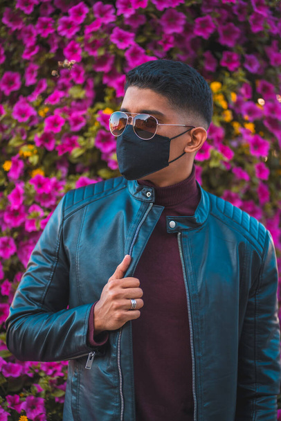 Fashion lifestyle, portrait of a young Latino with pink flowers in the background. Jeans, leather jacket and brown shoes. Portrait in the pandemic with mask - Zdjęcie, obraz