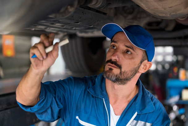 Mécanicien automobile en vêtements de travail inspecte une voiture dans un atelier d'une voiture. Un mécanicien de voiture tenant stylo. - Photo, image