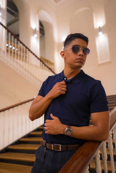 Fashion lifestyle, portrait of a young Latino man in a beautiful old building. Blue polo shirt and plaid pants. Portrait by the stairs - Fotó, kép