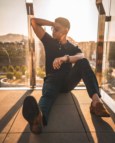 Fashion lifestyle, portrait of a young Latino with the city of San Sebastian in the background, Gipuzkoa. Blue polo shirt and plaid pants, sitting on the floor - Foto, immagini