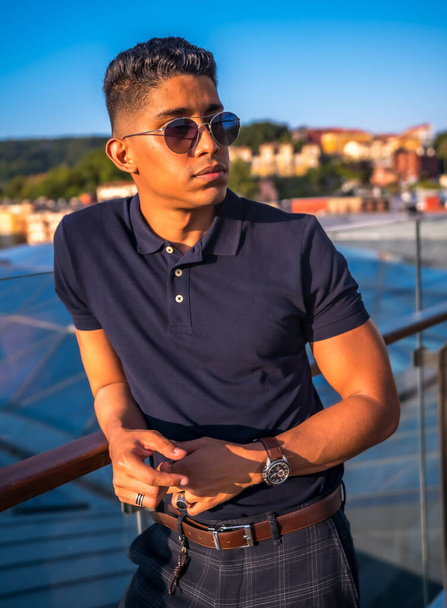 Fashion lifestyle, portrait of a young Latino with the city of San Sebastian in the background, Gipuzkoa. Blue polo shirt and plaid pants, pensive on the terrace - Φωτογραφία, εικόνα