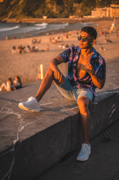 Summer lifestyle, portrait of a young Latino man enjoying summer vacation. with purple flower shirt and sunglasses, by the beach - Photo, Image