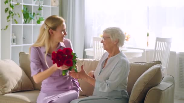 jong meisje geeft bloemen aan oud dame met vrolijke glimlach op verjaardag - Video