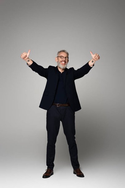full length of positive middle aged man in glasses and suit showing thumbs up on grey - Photo, Image