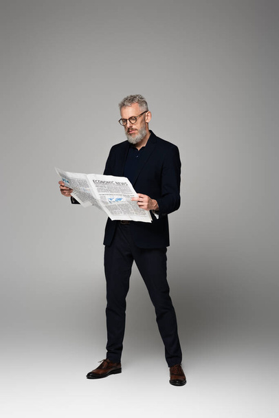 full length of middle aged man in glasses and suit reading economic news on grey - Foto, Bild