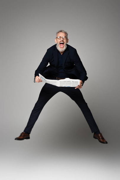 longitud completa de hombre sorprendido en gafas y traje celebración de periódico mientras levita en gris - Foto, imagen