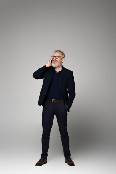 longitud completa de hombre feliz con el pelo gris hablando en el teléfono inteligente mientras posando con la mano en el bolsillo en gris - Foto, imagen