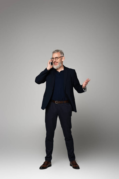 full length of happy man with grey hair talking on smartphone while gesturing on grey - Foto, imagen