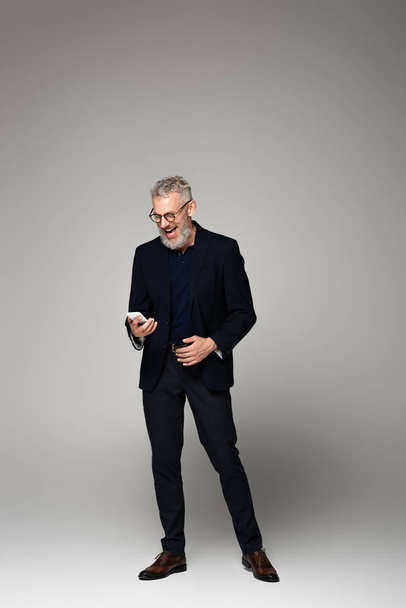 plná délka vzrušený podnikatel v obleku pomocí smartphone na šedé - Fotografie, Obrázek