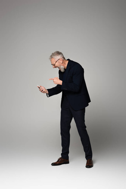 full length of happy businessman with grey hair pointing at smartphone on grey - Photo, Image