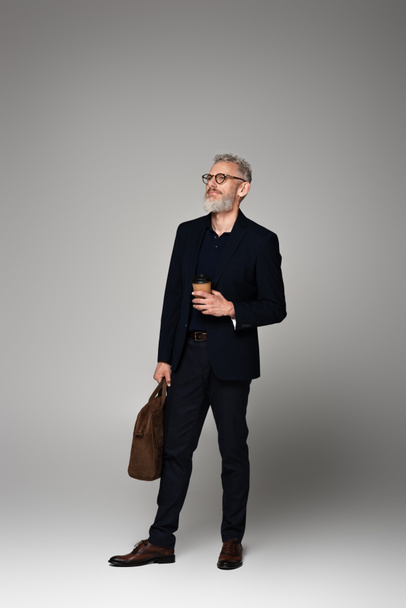 full length of man with grey hair in suit holding paper cup and leather bag on grey - Фото, изображение