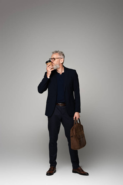 full length of man with grey hair in suit kissing paper cup and holding leather bag on grey - Foto, Imagen
