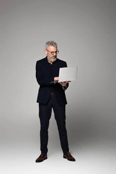 full length of man with grey hair in suit using laptop on grey - Foto, Bild