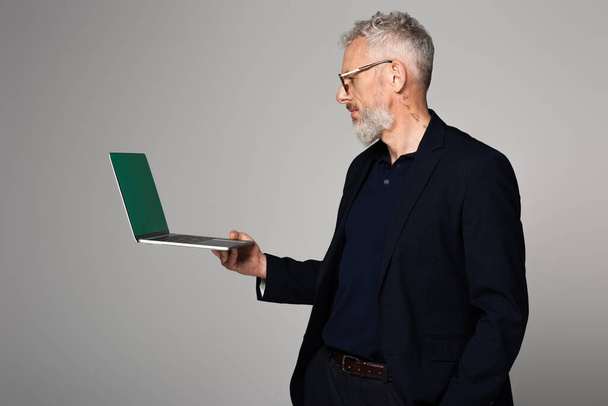getatoeëerde man van middelbare leeftijd in bril op zoek naar laptop geïsoleerd op grijs - Foto, afbeelding