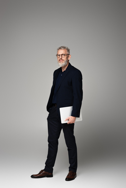 full length of man with grey hair in suit holding laptop while standing on grey - Foto, afbeelding