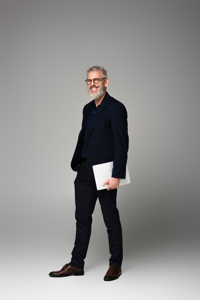 full length of happy man with grey hair holding laptop while standing on grey - Foto, afbeelding