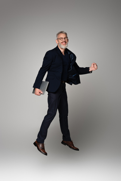 full length of cheerful man with grey hair holding laptop while levitating on grey - Foto, immagini