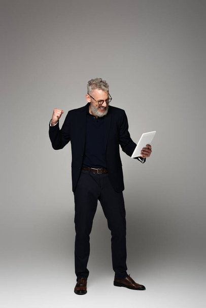 full length of excited middle aged businessman with grey hair looking at digital tablet and rejoicing on grey - Foto, afbeelding