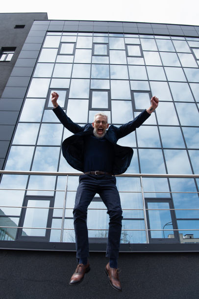 pełna długość dojrzałego mężczyzny z siwymi włosami w okularach skaczący w pobliżu nowoczesnego budynku na szarości  - Zdjęcie, obraz