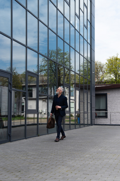 full length of businessman with grey hair holding leather bag and paper cup while walking near modern building  - Foto, imagen