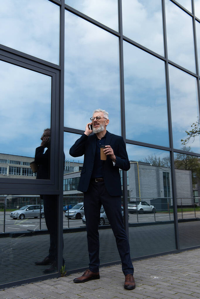 pełna długość dojrzały mężczyzna z siwymi włosami trzymając papierowy kubek podczas rozmowy na smartfonie w pobliżu nowoczesnego budynku  - Zdjęcie, obraz