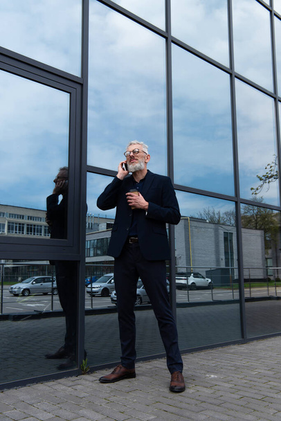 Geschäftsmann mit grauen Haaren hält Pappbecher in die Höhe, während er in der Nähe moderner Gebäude mit dem Smartphone spricht  - Foto, Bild