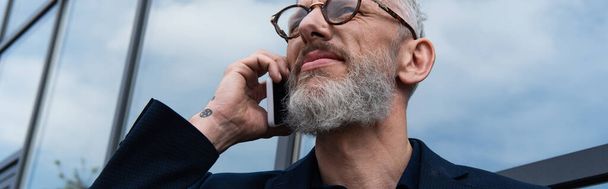 Gri saçlı olgun iş adamı modern binanın yakınında akıllı telefondan konuşuyor, afiş. - Fotoğraf, Görsel