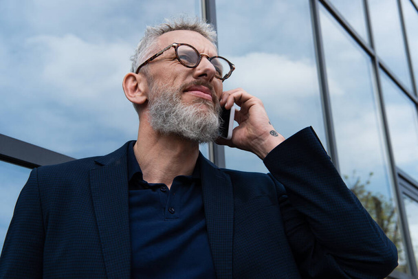 middle aged businessman in glasses with grey hair talking on smartphone near building - Photo, Image