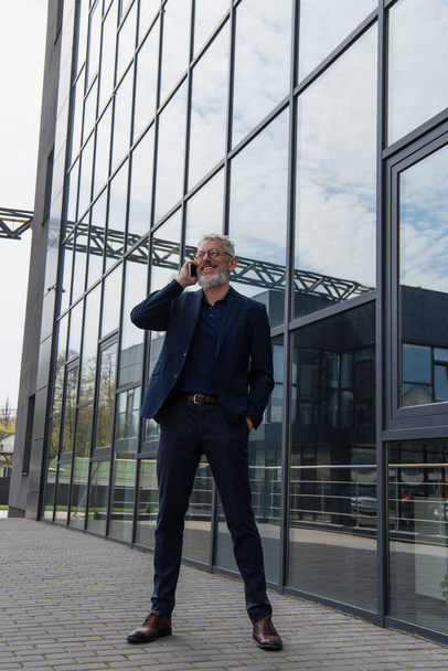 full length of happy middle aged businessman in glasses talking on smartphone near building - Photo, Image