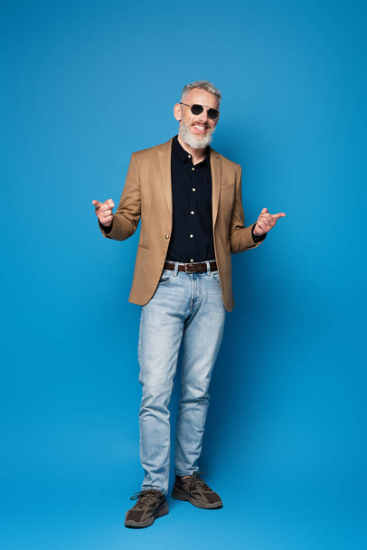 full length of happy middle aged man in sunglasses pointing with fingers on blue - Foto, afbeelding