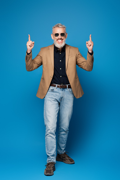 full length of cheerful middle aged man in sunglasses pointing up with fingers on blue - Zdjęcie, obraz